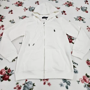 Vit hoodie från Ralph Lauren - Säljer en stilren vit hoodie från Ralph Lauren med dragkedja och huva. Tröjan har en liten broderad logga på bröstet och är tillverkad i mjuk bomull. Perfekt för en avslappnad look.