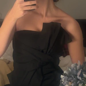 Svart topp - Snygg svart axelbandslös tooo med en elegant drapering över bysten. Perfekt för en kväll ute eller en speciell tillställning. Toppen har en stilren och modern design som framhäver axlarna.