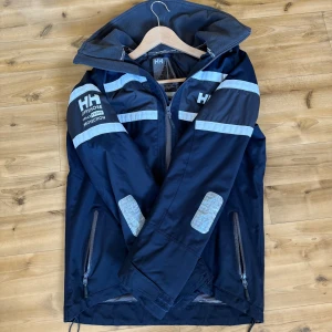 Marinblå jacka från Helly Hansen - Säljer en marinblå jacka från Helly Hansen med dragkedja och huva. Jackan har vita detaljer och reflexband på ärmar och bröst. Perfekt för utomhusaktiviteter med sin robusta design och skyddande material.