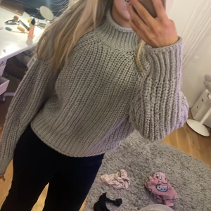 Grå stickad tröja från H&M - Mysig grå stickad tröja från H&M med en hög krage och långa ärmar. Perfekt för kyliga dagar och ger en avslappnad look. Tröjan har en ribbad struktur som ger extra stil och komfort.