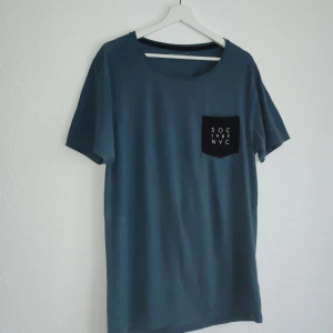 Blå t-shirt med bröstficka från SOC - Säljer en blå t-shirt från SOC med en stilren bröstficka där det står 'SOC 1989 NYC'. T-shirten har en avslappnad passform och korta ärmar, perfekt för en casual look.