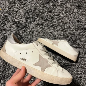 Vita sneakers från Golden Goose - Snygga vita sneakers från Golden Goose med en ikonisk stjärndetalj på sidan. Skorna har en vintage-look med grå snörning och en smutsig finish för en avslappnad stil. Skorna är använda, men funkar fortfarande och därför är priset mycket bra. Den ända defekten skorna har är att det fattas två bokstäver på sidan av skon.