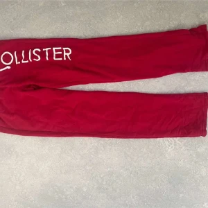 Röda mjukisbyxor från Hollister - Snygga röda mjukisbyxor från Hollister med vit logga och siffrorna '1922' tryckta på sidan. Byxorna har en avslappnad passform och är perfekta för en chill dag hemma. De har elastiska muddar vid bensluten för extra komfort.
