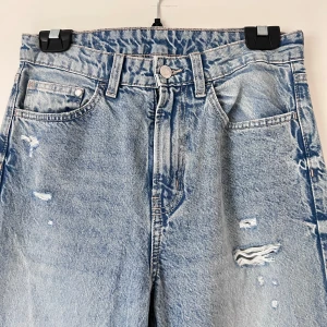 Blå jeans med slitningar - Snygga blå jeans med slitningar och en avslappnad passform. De har en klassisk femficksdesign och en knappgylf. Perfekta för en trendig och avslappnad stil. Använda runt 2 gånger. Önskar du veta exakta mått? Tveka inte att höra av dig!