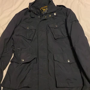 Woolrich Field jacka - Säljer en svart jacka från Woolrich med flera fickor och knappar. Jackan har en stilren design med långa ärmar och är perfekt för kyligare väder. Den har en praktisk dragkedja och knappar för extra skydd mot vind. En knapp är borta vid fickan men det syns inte. Pris är inte hugget i sten 