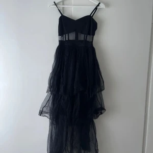 Svart långklänning med tyll - Snygg svart långklänning med tunna axelband och transparenta detaljer i midjan. Kjolen är i flera lager av tyll som ger en voluminös look. Perfekt för en elegant kväll.