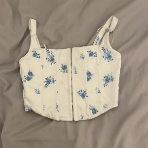 Vit blommig korsett från Gina Tricot - Säljer en söt vit korsett från Gina Tricot med blått blommönster. Den har hakknäppning framtill. 🌸