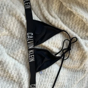 Svart bikini från Calvin Klein - Snygg svart bikini från Calvin Klein med märkets logga på banden. Toppen har en trekantsform och knytning i nacken. Perfekt för en stilren look på stranden eller vid poolen. Den är äkta är köpt på Calvin Klein hemsida. Inget fel på den den är lite använd. Den har inga inlägg i sig dom är borta men har man några egna inlägg så kan man sätta i de om man vill. Priset är inte satt är öppen för alla pris förslag 