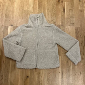 Beige teddyjacka från H&M - Mysig beige teddyjacka från H&M med dragkedja framtill. Jackan har en hög krage och två stora fickor.