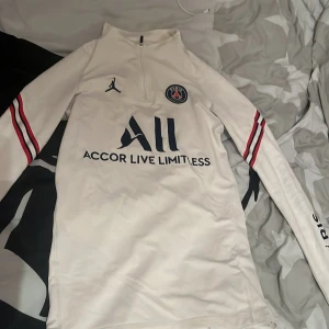 Vit långärmad tröja från Jordan x PSG - Säljer en vit långärmad tröja från Jordan x PSG med dragkedja vid halsen. Tröjan har röda och blå ränder på ärmarna och texten 'Accor Live Limitless' och 'Visit Rwanda'. Perfekt för fotbollsfans!