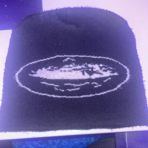 Svart mössa med vit logga - Snygg svart mössa med en vit broderad logga framtill. Perfekt för att hålla värmen under kyliga dagar. Enkel och stilren design som passar till det mesta. Säljs pga tuffa tider för plånboken😅