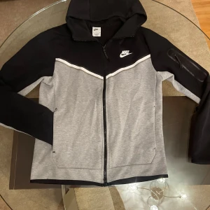 Grå och svart nike tech - Säljer en snygg grå och svart jacka från Nike med dragkedja och huva. Jackan har en praktisk ficka med dragkedja på ärmen och ett stilrent Nike-logotyp på bröstet. Perfekt för en sportig look!