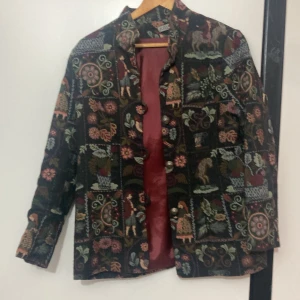 Mönstrad jacka från Kriss - Säljer en unik jacka från Kriss med ett färgglatt och detaljerat mönster som inkluderar blommor och figurer. Jackan har långa ärmar och knappar framtill. Perfekt för dig som vill sticka ut med en konstnärlig stil.