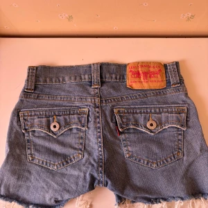 Blå jeansshorts från Levi's - Säljer ett par snygga blå lågmidjade jeansshorts från Levi's med fransiga kanter och slitna detaljer. Shortsen har klassiska femficksdesign och en knappgylf. Perfekta för en avslappnad stil.