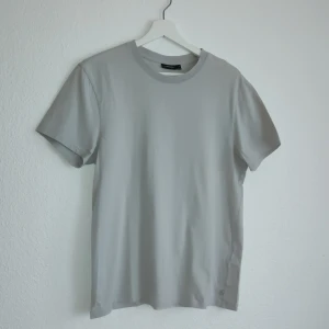 Grå t-shirt från J.Lindeberg - Säljer en stilren grå t-shirt från J.Lindeberg. Den har en klassisk rund hals och korta ärmar, perfekt för en minimalistisk look. Tillverkad i mjukt material för en bekväm passform.