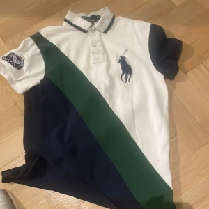 Vit och marinblå pikétröja från Ralph Lauren - Säljer en snygg pikétröja från Ralph Lauren i vitt och marinblått med en grön diagonal rand. Tröjan har korta ärmar och en broderad logga på bröstet samt ett Wimbledon-märke på ärmen. Perfekt för en sportig look!