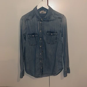 Blå jeansskjorta med knappar - Blå jeansskjorta med långa ärmar och knappar framtill. Skjortan har två bröstfickor med knappdetaljer och en klassisk krage. 