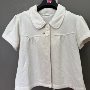 Vit kortärmad kofta med krage - Säljer en söt vit kofta med korta ärmar och en fin krage. Koftan har två knappar framtill och en lätt rynkad detalj under bysten. Perfekt för en stilren och bekväm look.