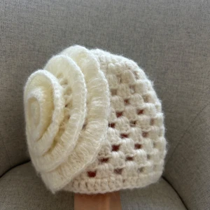 Vit stickad mössa med blomdetalj - Charmig vit stickad mössa med en stor blomdetalj på sidan. Perfekt för att hålla värmen med stil under kyliga dagar. Den har ett luftigt mönster som ger en unik look.