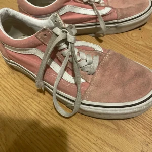 Rosa och vita sneakers från Vans - Säljer ett par klassiska rosa och vita Vans sneakers med snörning. Skorna har en ikonisk vit sidosöm och är tillverkade i canvas med mockadetaljer. Perfekta för en avslappnad stil. lite smutsig men annars helt fungerande