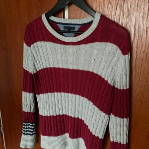 Randig stickad tröja från Tommy Hilfiger - Säljer en snygg stickad tröja från Tommy Hilfiger med breda vinröda och grå ränder. Tröjan har rund halsringning och långa ärmar, samt diskret logga på bröstet. Perfekt för dig som gillar klassisk stil med en twist. Funkar toppen till sommaren. Pris kan diskuteras vid snabb affär!