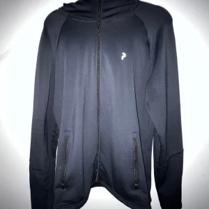 Svart hoodiejacka från peak performance  - Svart hoodiejacka med dragkedja från peak performance . Jackan har två fickor med dragkedja framtill, liten vit logga på bröstet och huva med tryckt text. Perfekt för en sportig och stilren look.