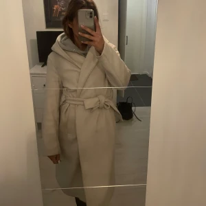 Beige kappa med huva och bälte - Säljer en beige kappa med huva och knytskärp i midjan. Jackan har lång ärm och en klassisk omlottmodell som ger en stilren look. Perfekt för kyligare dagar när du vill vara både varm och snygg.