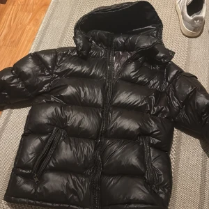 Svart dunjacka från Moncler - Säljer en svart pufferjacka från Moncler med huva och dragkedja. Jackan har två stora fickor med dragkedja framtill och Moncler-logga på ena ärmen. Perfekt för kyliga dagar .   finns en liten skada som ni ser på sista bilden, kom med bud