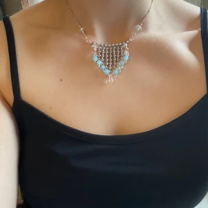 Silverfärgat halsband med blå pärlor - Unikt halsband i silverfärg med flera rader av små kulor och blå pärlor. Hängande design med droppformad detalj i mitten. Perfekt för att ge en färgklick till din outfit.