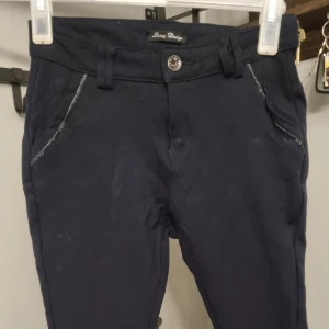 Mörkblå kostymbyxor från Shiny Design - Mörkblå kostymbyxor med normal passform från Shiny Design. Byxorna har snedställda fickor fram och dekorativa fickor bak. Klassisk knapp och hällor för bälte i midjan. Perfekta för en stilren look.