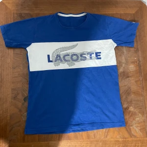 Blå t-shirt från Lacoste med logga - Säljer en blå t-shirt från Lacoste med vit panel och stor logga framtill. Klassisk rund hals och korta ärmar. Perfekt för en sportig och avslappnad stil.