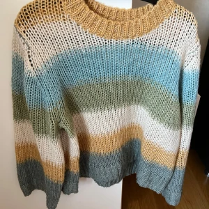 Randig stickad tröja - Säljer en stickad tröja från Lindex med breda ränder i blått, grönt, vitt, beige och gult. Tröjan har grovstickad struktur, rund halsringning och långa ärmar. Perfekt för dig som gillar färg och mysiga plagg!
