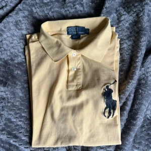 Gul piké från Polo Ralph Lauren - Snygg gul pikétröja från Polo Ralph Lauren med klassisk krage och två knappar. Stor svart broderad logga på bröstet och siffra på ärmen. Perfekt för en stilren och sportig look. 