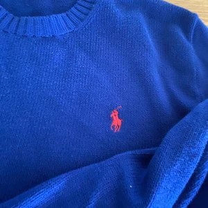 Blå stickad tröja från Polo Ralph Lauren - Säljer en klassisk blå stickad tröja från Polo Ralph Lauren med rund halsringning och röd broderad logga på bröstet. Perfekt till jeans eller chinos för en stilren look.
