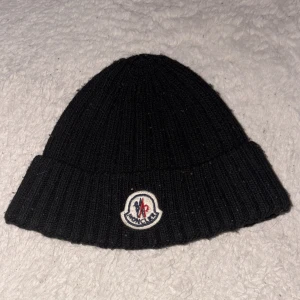 Svart ribbstickad mössa från Moncler - Svart ribbstickad mössa från Moncler med uppvikt kant och klassisk Moncler-logga framtill. Perfekt för kyliga dagar och enkel att matcha med olika outfits. Materialet känns mjukt och värmande.