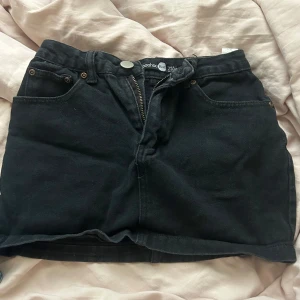 Svart jeanskjol från Boohoo - Svart kort jeanskjol från Boohoo med klassisk femficksdesign och dragkedja framtill. Kjolen har bälteshällor och rå kant nertill 