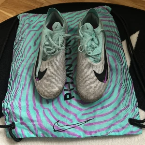 Nike Phantom GT fotbollsskor i mintgrön och grå - Säljer ett par Nike Phantom GT fotbollsskor i mintgrönt och grått med svarta och lila detaljer. Skorna har snörning och en platt sula med dobbar för bra grepp på planen. Perfekta för dig som vill sticka ut på fotbollsplanen! Dom är i bra skick men bara lite smutsiga