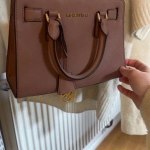 Brun handväska från Michael Kors - Säljer en snygg brun handväska från Michael Kors med guldfärgade metalldetaljer och avtagbar axelrem. Väskan har dubbla handtag och ett innerfack med dragkedja. Perfekt för att bära dina viktigaste saker med stil. Använd ett fåtal gångeg därför inga defekter. Skriv för fler frågor eller bilder.🥰🥰