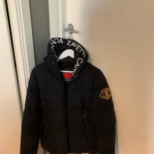 Svart pufferjacka från Zavetti Canada - Säljer en svart pufferjacka från Zavetti Canada med huva och broderad logga på ärmen. Jackan har dragkedja framtill och två dragkedjefickor. Fodrad och perfekt för kyligare dagar.