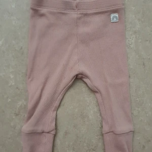 Leggings - Supersöta ljusrosa ribbade leggings från Lindex Baby. Byxorna har en mjuk resår i midjan och en liten tygpatch med regnbågsmotiv framtill. Perfekta för små barn som vill ha något bekvämt och mjukt på sig.