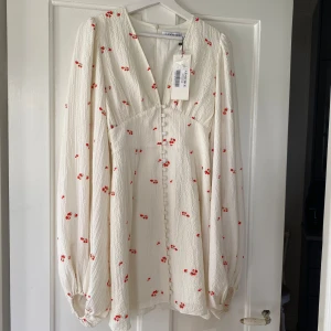 Vit blommig klänning från Adoore - Superfin vit klänning från Adoore med små röda blommor och v-ringning. Klänningen har långa puffärmar och knappar framtill. Perfekt för vår och sommar!🩷Aldrig använd!