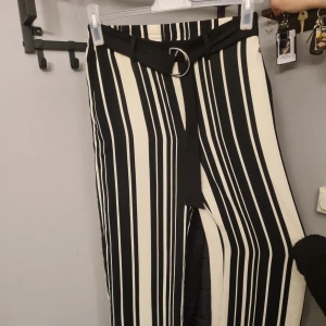 Svartvita randiga byxor med bälte från River Island - Snygga svartvita byxor från River Island med vertikala ränder och tillhörande svart bälte med metallspänne. Byxorna har en rak passform och elastisk midja baktill för extra komfort. Perfekta för dig som vill sticka ut med en trendig look.