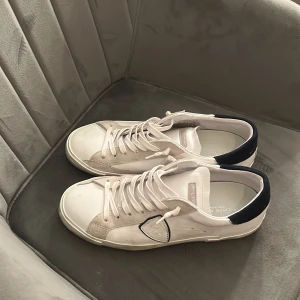 Vita sneakers från Philippe Model - Säljer ett par vita Philippe Model sneakers med mörkblå detaljer på hälen och gråa partier vid snörningen. Knappt använda. Pris kan diskuteras. Har tyvärr inte kvar Phillippe Model Boxen.