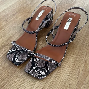 Ormskinnsmönstrade sandaler med klack - Säljer ett par trendiga sandaler med ormskinnsmönster i svart, grått och beige. Skorna har fyrkantig tå, smala remmar och en stabil klack. 