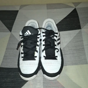 Vita och svarta sneakers från Adidas - Säljer ett par klassiska vita sneakers från Adidas med svarta detaljer och tre ränder på sidan. Skorna har snörning, perforerad ovandel och platt sula. Perfekta för dig som gillar sportig stil.