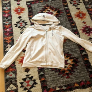 Vit hoodie från Sail Racing med dragkedja - Säljer en vit hoodie från Sail Racing med dragkedja framtill och en extra dragkedjeficka på bröstet. Jackan har huva med broderad logga och mjukt fleecefoder på insidan. Perfekt för dig som gillar sportig stil.