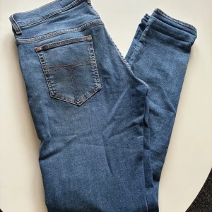 Tiger of Sweden Jeans - Säljer ett par helt nya klassiska blå jeansbyxor ifrån Tiger of Sweden med fem fickor och orangea sömmar. Byxorna har normal passform och raka ben, perfekt för en enkel och stilren look. Bälteshällor och knappgylf upptill. Ny pris: 1599kr    Strl: W30/L32