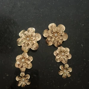 Guldiga blommiga örhängen med pärldetaljer - Säljer ett par stora statement-örhängen i guld med tre blommor i olika storlekar på varje örhänge. Varje blomma har en liten pärla i mitten som detalj. Perfekta för att lyfta vilken outfit som helst!