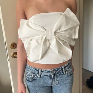 Vit tubtopp med stor rosett från Kappahl - Supersöt vit tubtopp från Kappahl med en stor rosett framtill. Toppen är axelbandslös/bandau och är aldrig använd🤍