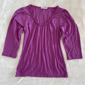 Lila topp med volangkant från Wera - Säljer en lila topp från Wera med volangkant runt den djupa v-ringningen. Toppen har trekvartsärm och är gjord i ett mjukt material, perfekt för en färgglad vardagslook.